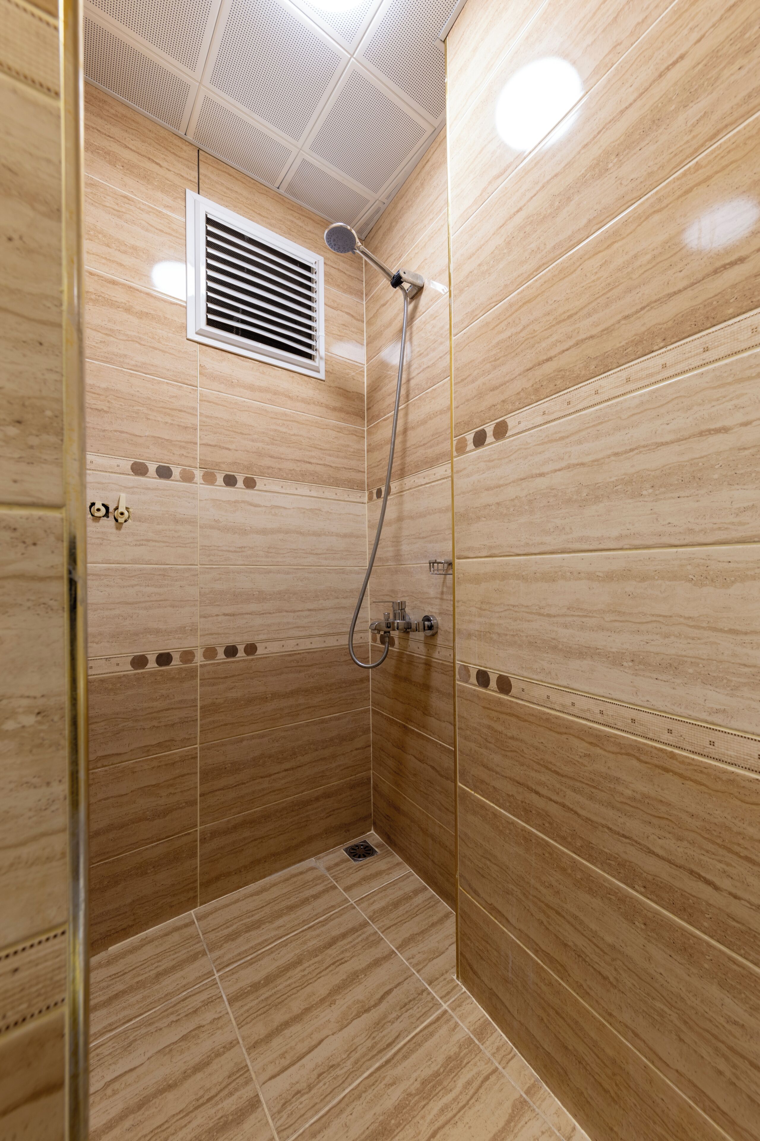 Een douche met beige tegels, ventilatie en een handdouche.
