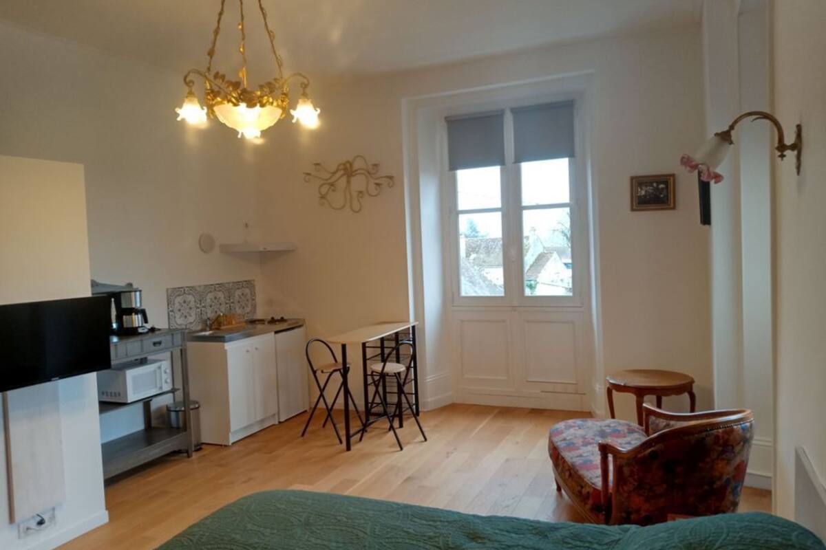 A room with a bed, a desk, a chair, a small table, a window, and a kitchenette.