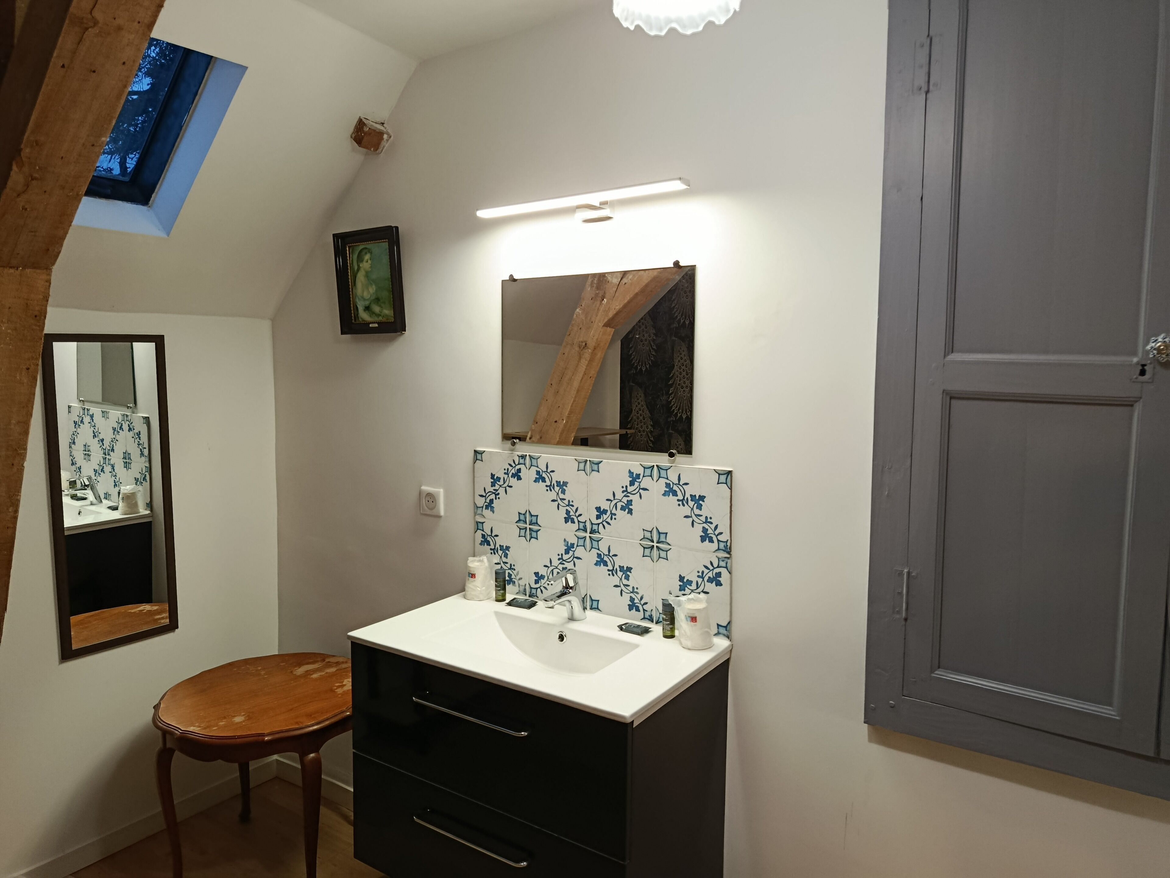 Un baño con lavamanos blanco, mueble negro, taburete de madera y un espejo.