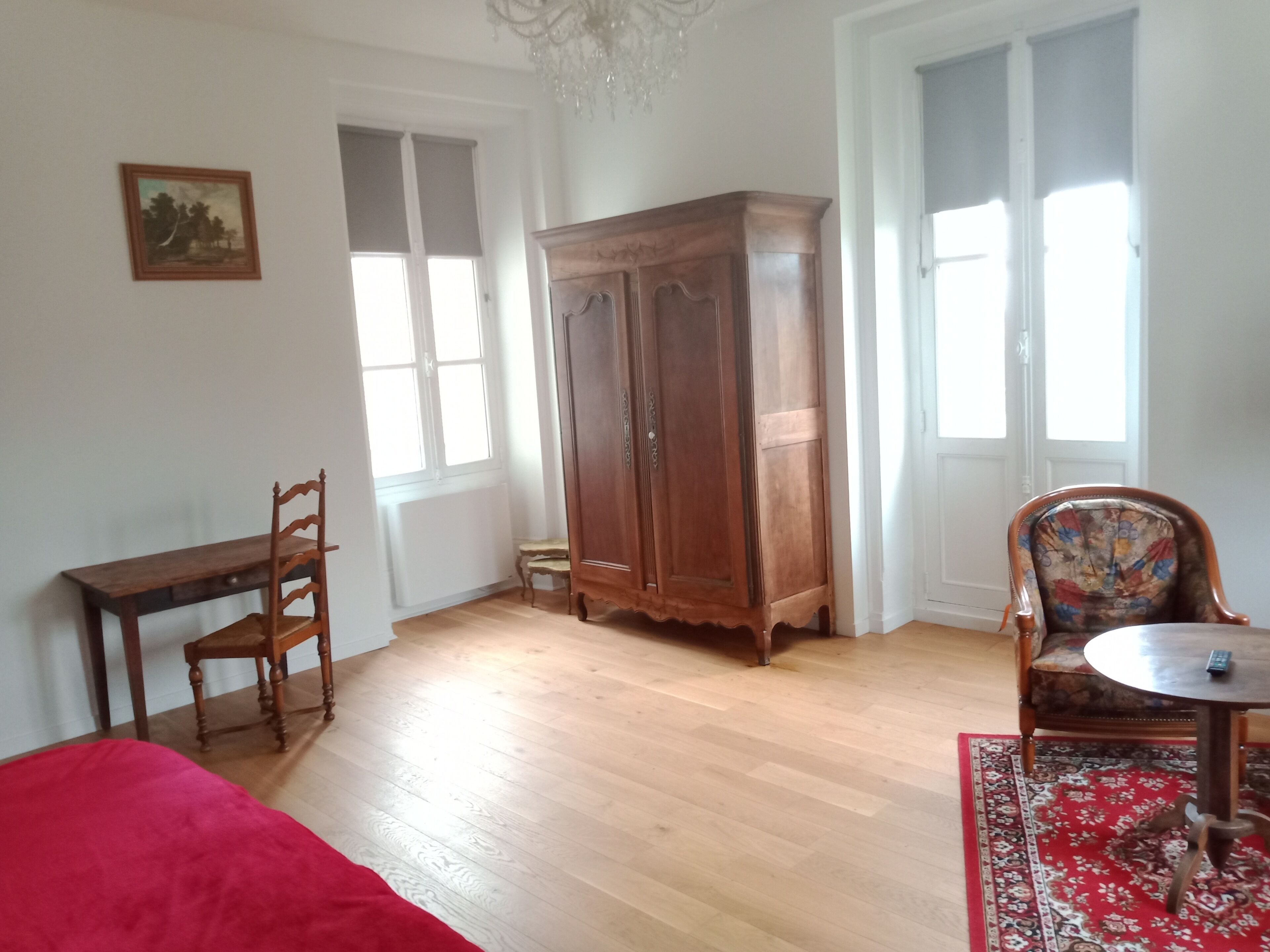 A room with a wooden floor, a red bed, a wooden desk with a chair, a large wooden wardrobe, a patterned armchair, and a small table with a remote control.