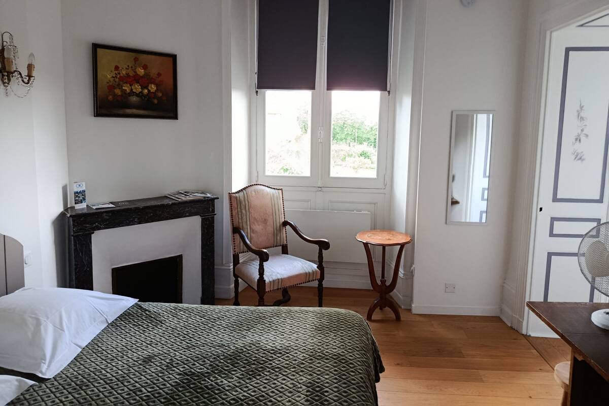 A room with a bed, a chair, a small table, a fireplace, a window with blinds, and a door.