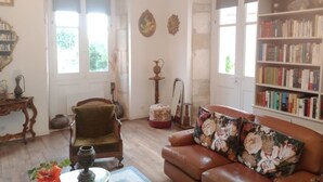 A cozy living room with a leather sofa, a bookshelf, a wooden chair, and a large window. - VILLA SAINT-PIERRE (La Trimouille)