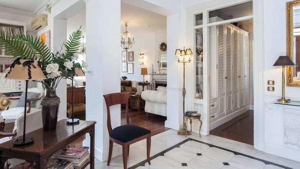 Una habitación con una silla de madera, un florero con flores, una lámpara, una estantería y una puerta que da a otra habitación.