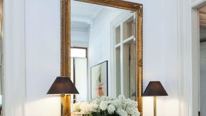Pared blanca con un espejo grande, dos lámparas y un florero con flores blancas sobre una consola de vidrio.