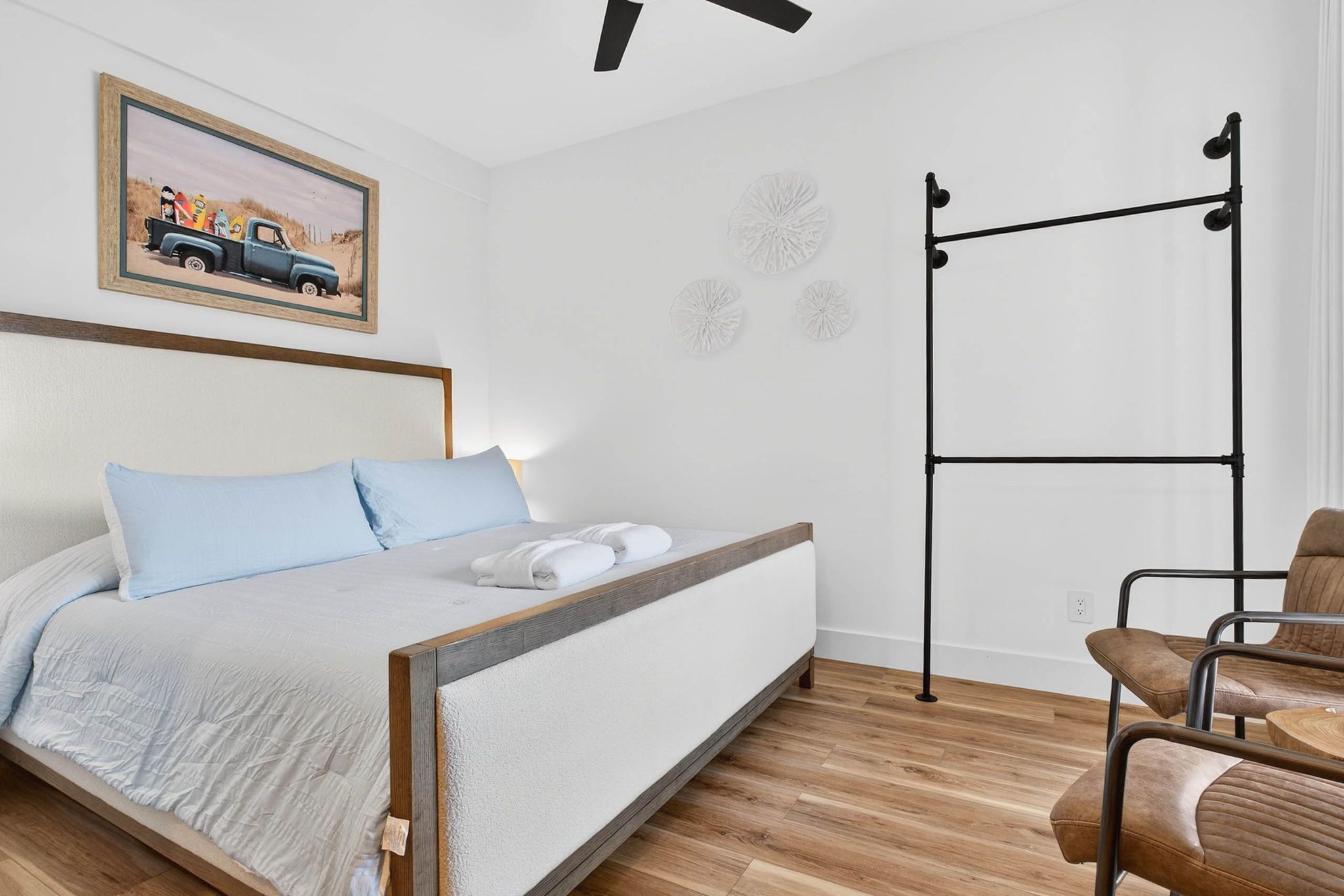 A bed with a headboard, a painting of a truck on the wall, a ceiling fan, and a metal rack against the wall.