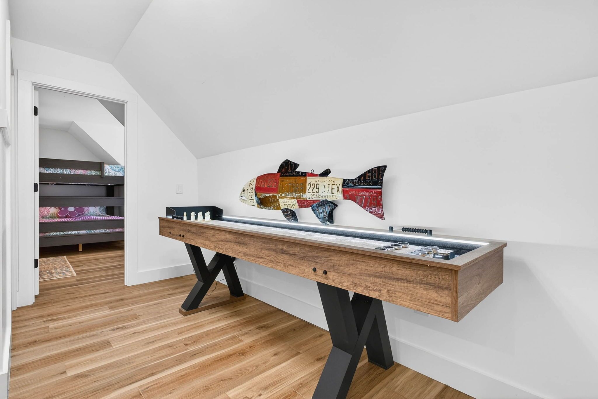 A room with a long wooden table, a wall-mounted fish decoration, and a staircase leading to a bed with patterned bedding.