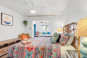 A bedroom with a bed, a bedside table, a lamp, a ceiling fan, a window, and a door leading to another room.