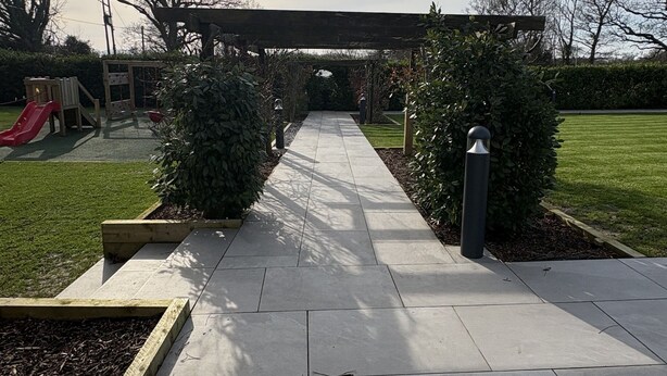 A paved pathway leading to a wooden pergola with a slide and climbing frame, surrounded by greenery and a clear sky.