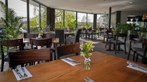 A restaurant with wooden tables and chairs, large windows, and indoor plants.