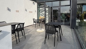 An outdoor patio with tables and chairs, a glass door entrance, and a white wall with a 'BAR' sign.