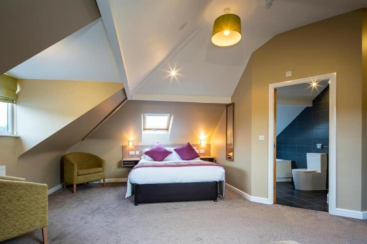 A bedroom with a bed, a chair, a nightstand, a wall-mounted lamp, a ceiling light, and a view into an adjacent bathroom.