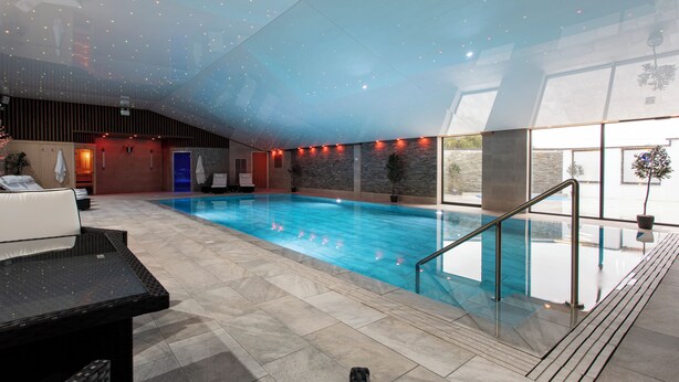 An indoor swimming pool with a glass ceiling, a lounge area with seating, and a view of the outdoors.