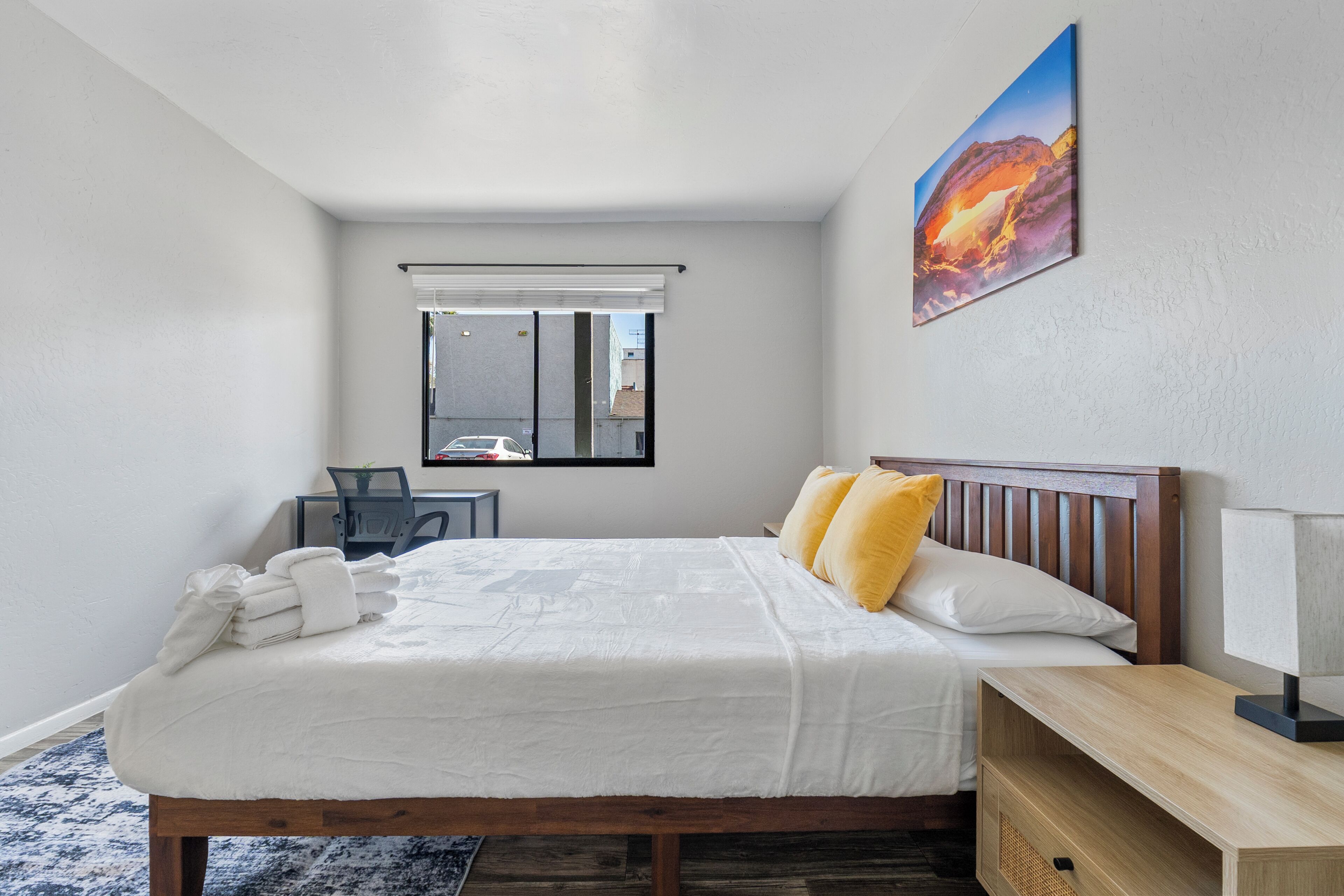 Een slaapkamer met een bed, nachtkastje, lamp en een schilderij aan de muur.