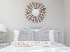 A bed with a white headboard, a decorative mirror, and a pillow with the text 'LIFE IS BETTER AT THE BEACH'. - Brass Rail 310 (Tybee Island)