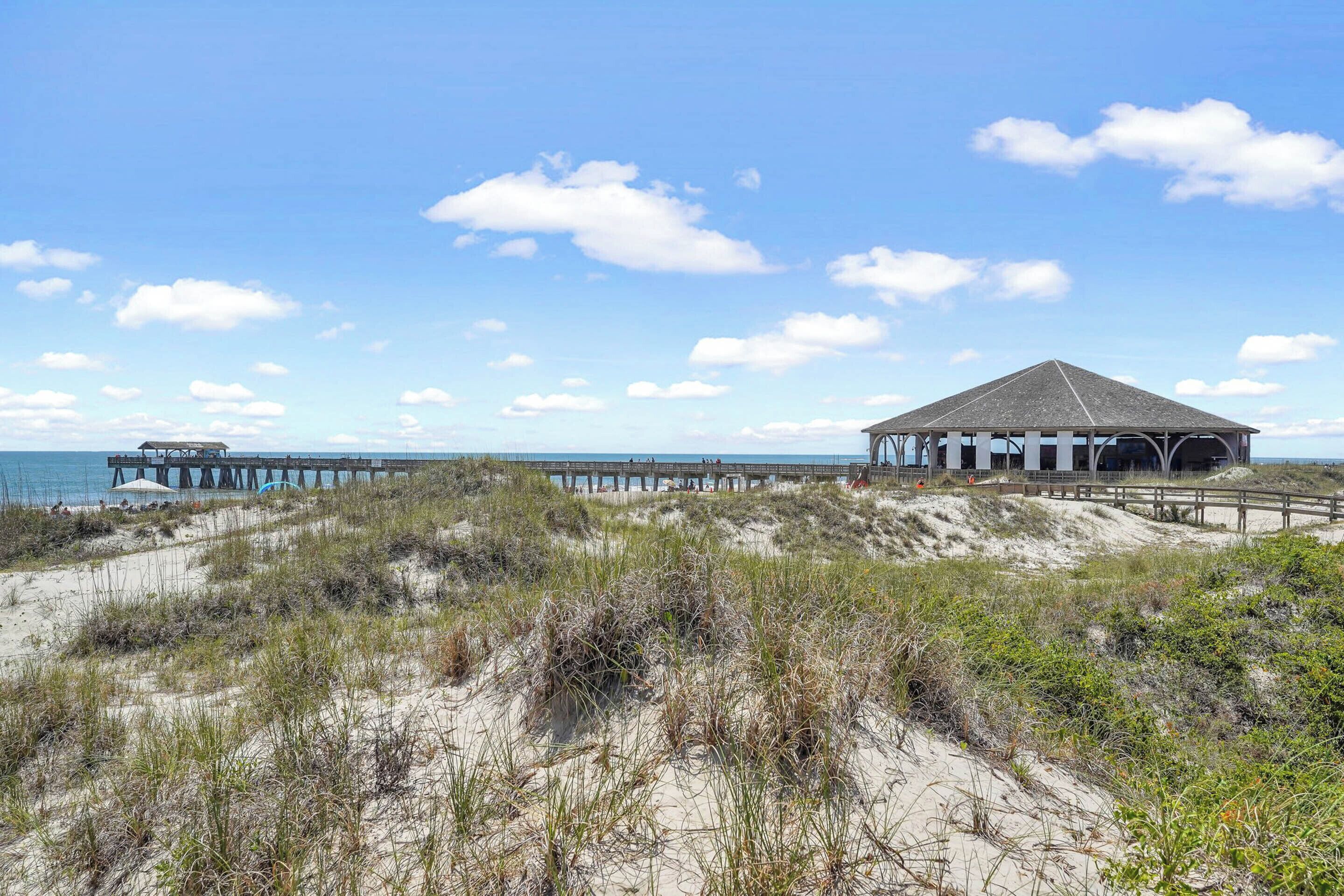 A beach with a wooden pier extending into the ocean, a covered structure, and sandy dunes with sparse vegetation.