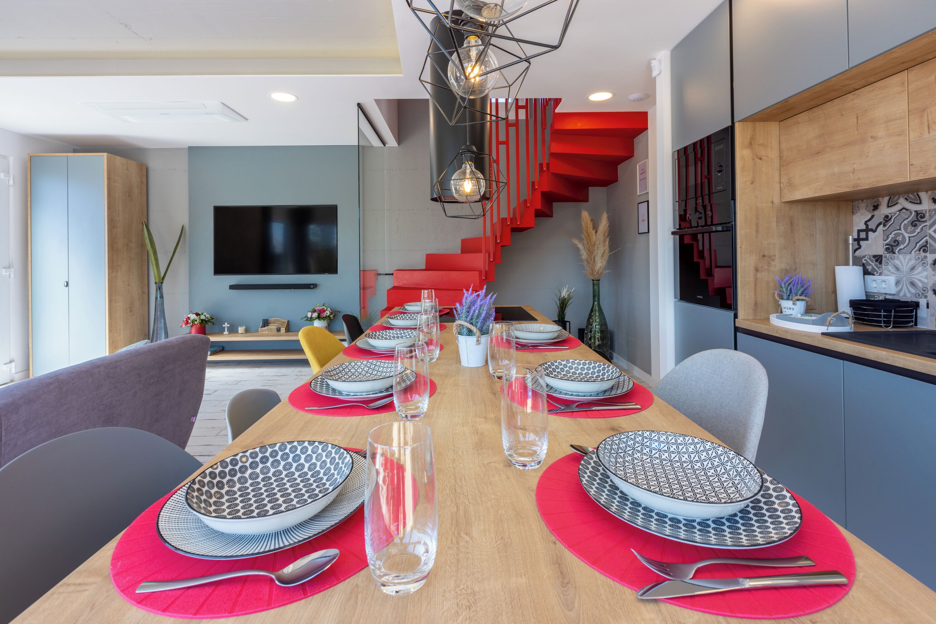 A modern dining area with a long table set for six, a red staircase, and a kitchen area with wooden cabinets.