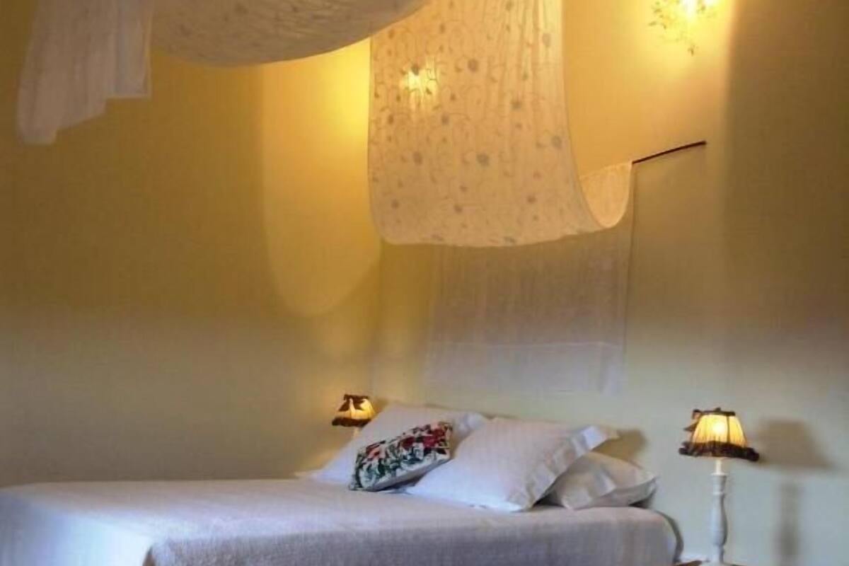 A bedroom with a bed, a bedside table with a lamp, a hanging canopy, and a wall-mounted light fixture.