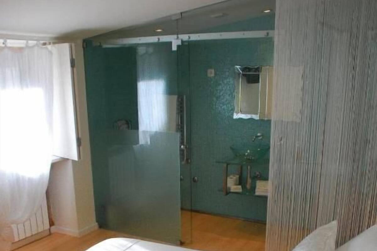 A bedroom with a bed, a bathroom with a shower, and a glass door leading to another room.