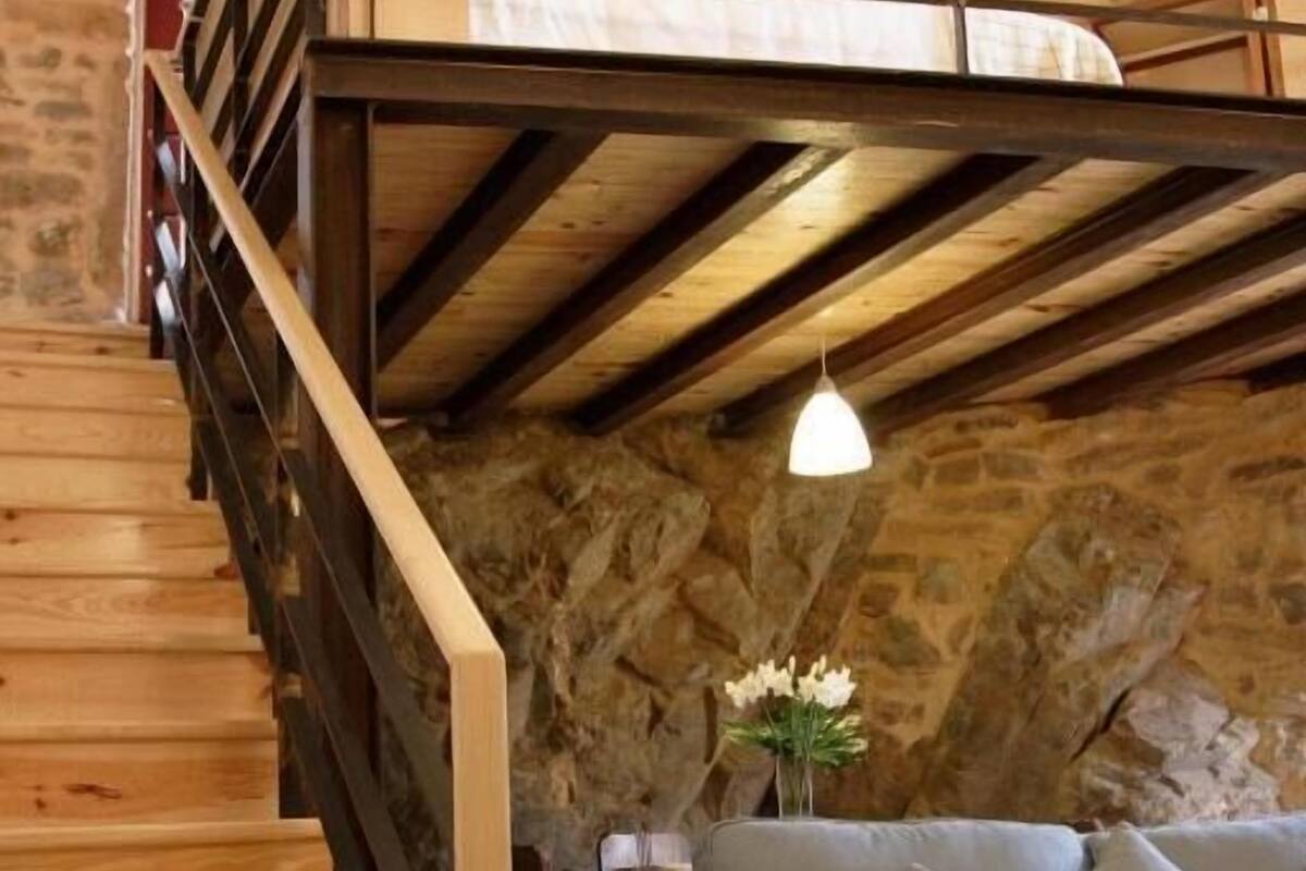 A wooden staircase leading to a loft bedroom with a stone wall, a gray sofa, and a hanging light fixture.