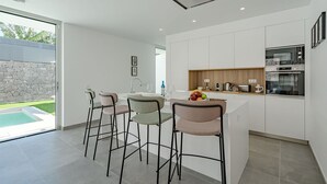 A modern kitchen with white cabinets, a built-in oven, and a dining area with bar stools. - Standard Villa 2 - Casangular by Upper (Adeje)