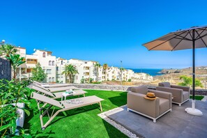 Eine Terrasse mit Gartenmöbeln, einem Tisch und einem Sonnenschirm mit Blick auf die Küste, Gebäude und das Meer.