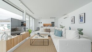 A modern living room with a sofa, coffee table, and TV, a kitchen in the background, and large windows. - Standard Villa 6 - Casangular by Upper (Adeje)
