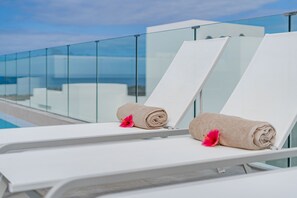 Zwei weiße Liegestühle mit Handtüchern und rosafarbenen Blumen, mit Blick auf einen Pool und einen wolkenlosen Himmel.