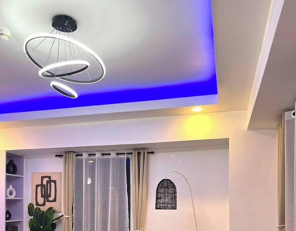 A modern interior with a ceiling light fixture, a window with curtains, and a wall-mounted shelf with decorative items. - Welcome to Lucky7suites (Parañaque)