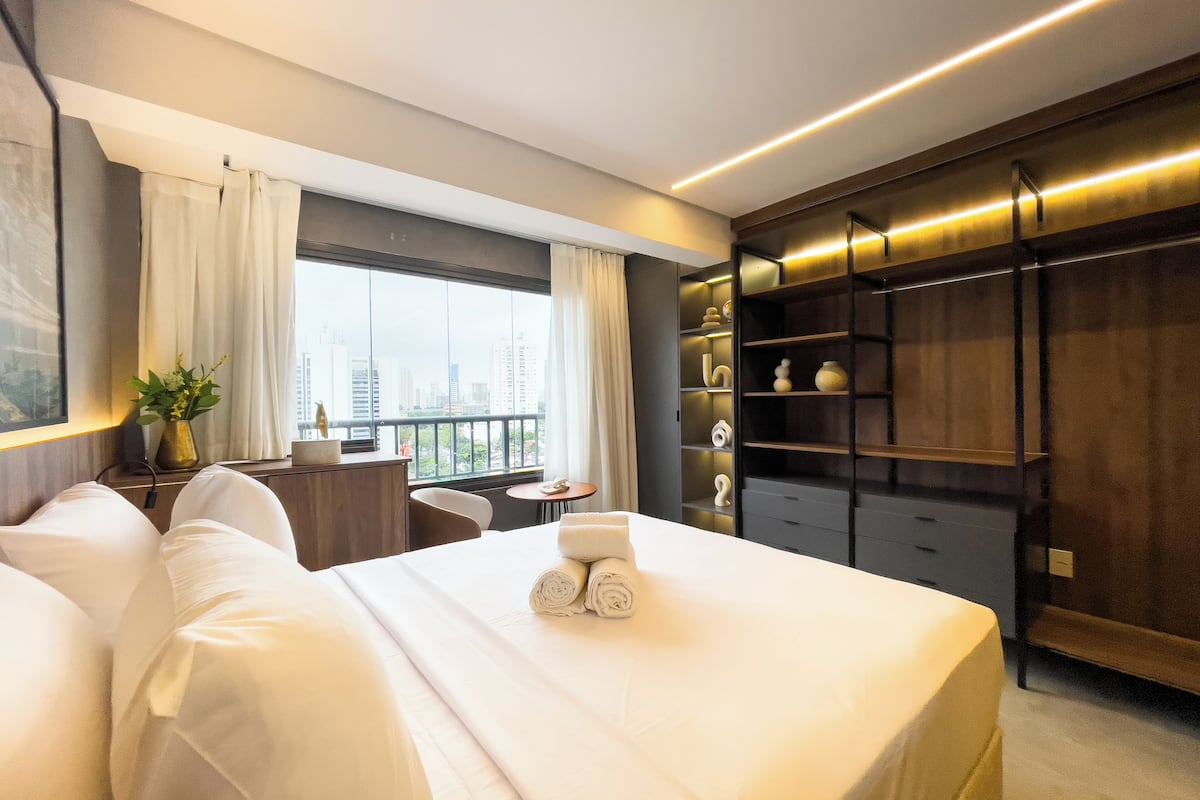 Habitación de hotel con una cama grande, un escritorio, una silla, una mesita, una ventana con cortinas y un armario empotrado con estantes.