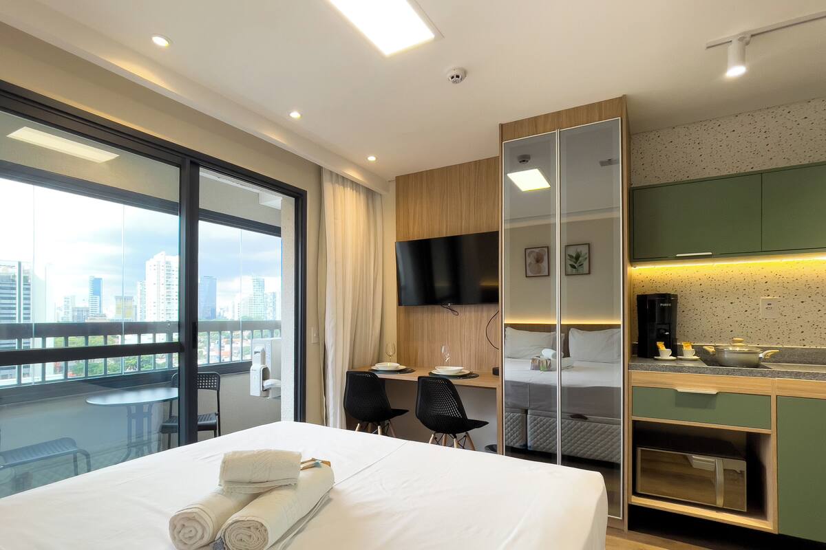 Habitación de hotel con cama, escritorio, silla, televisor, kitchenette y vista a la ciudad.