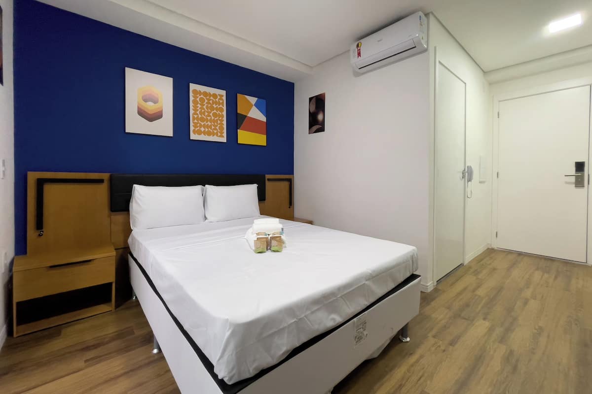 Una habitación de hotel con cama, mesita de noche, aire acondicionado instalado en la pared y una puerta.