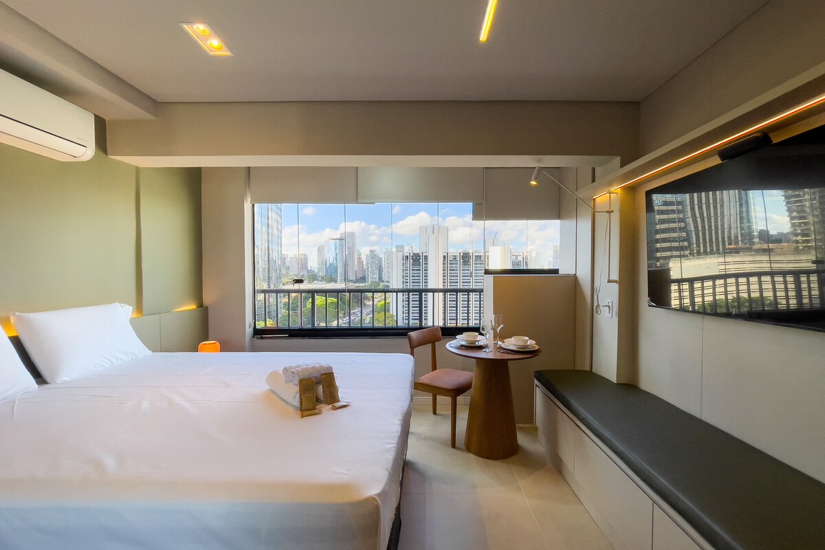 Habitación de hotel moderna con una cama grande, un área de comedor con mesa y sillas, y vista a la ciudad por la ventana.