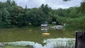 Une plage de sable avec un poteau en bois, un radeau jaune sur l’eau et une maison en arrière-plan.