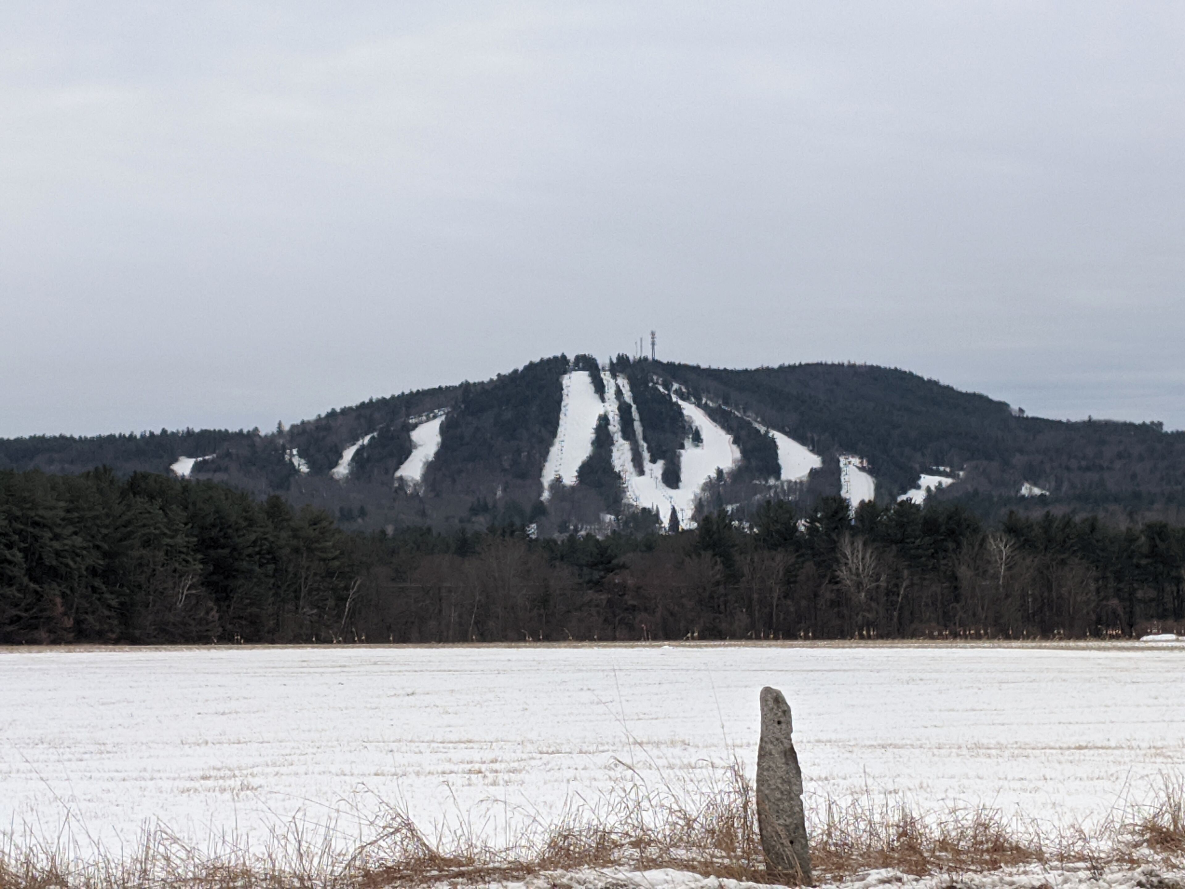 A snow-covered field with a prominent rock in the foreground and a forested hill in the background.