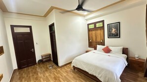 Une chambre à coucher avec un lit, un ventilateur de plafond, un plancher en bois, une porte et une étagère en bois sur laquelle est posée une image.