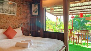 Une chambre d’hôtel avec un mur en briques, un lit recouvert de draps blancs et agrémenté d’oreillers rouges, une fenêtre donnant sur un paysage verdoyant et un luminaire suspendu.