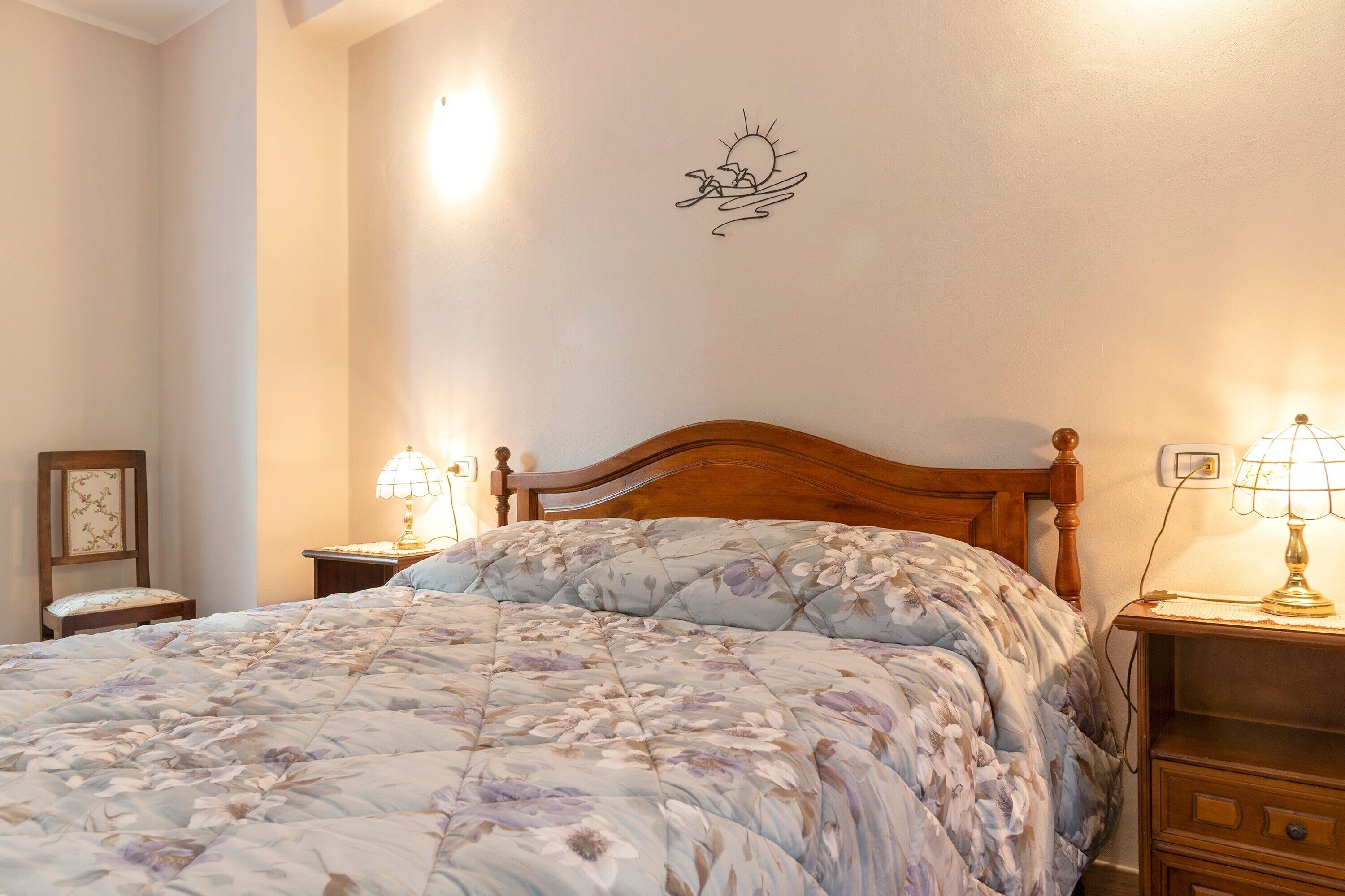 A bedroom with a large bed, two bedside tables with lamps, a wooden headboard, a wall decoration, and a small chair.