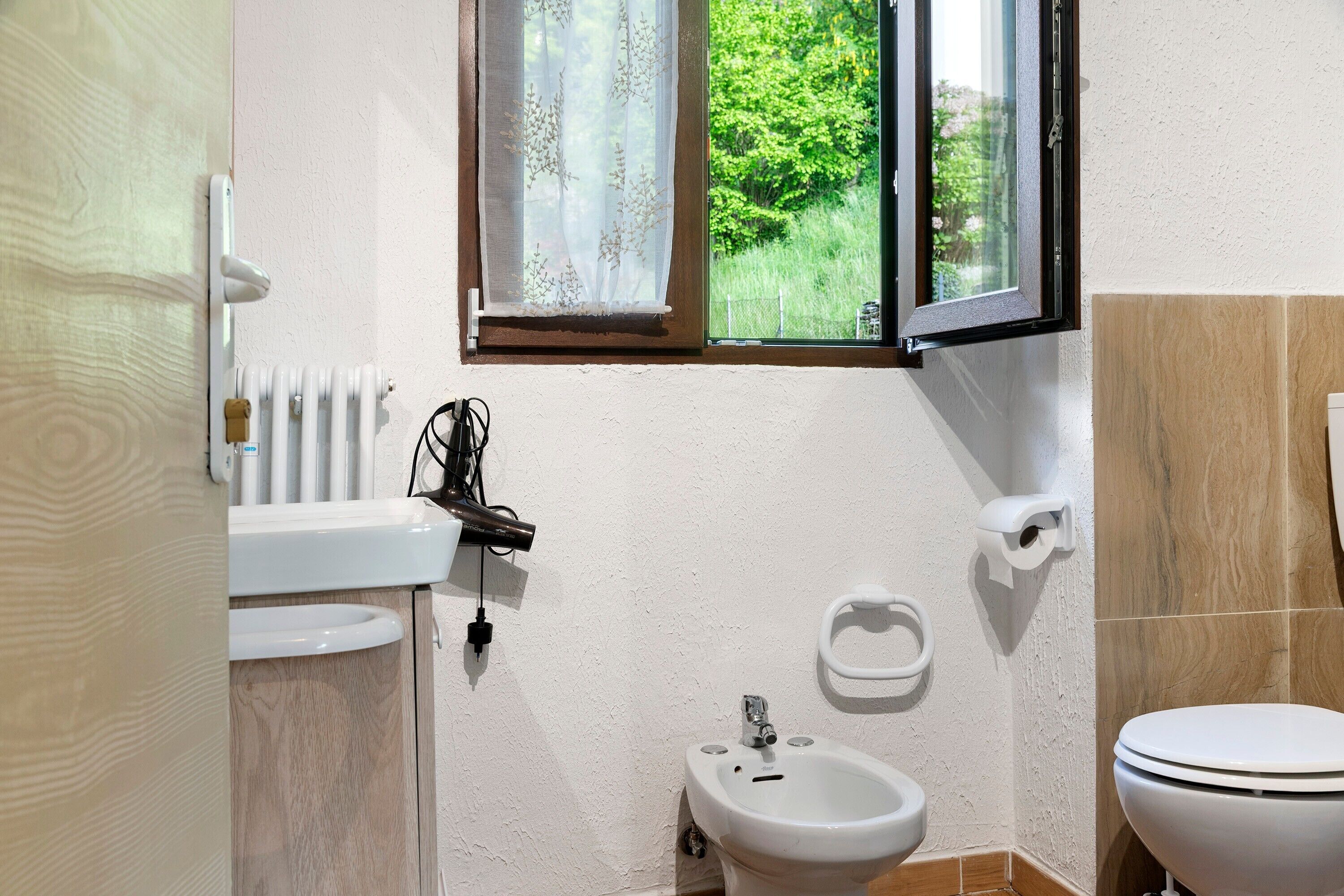 A bathroom with a toilet, bidet, and sink, a window with a view of greenery, and a radiator.