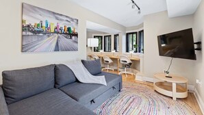 Ein modernes Wohnzimmer mit grauem Sofa, farbenfrohem Teppich, Flachbildfernseher und Stadtbildgemälde an der Wand.