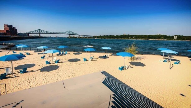 Ein Strand mit blauen Sonnenschirmen und Liegestühlen, ein Brücke im Hintergrund und ein klarer Himmel.