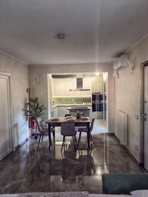 Une cuisine moderne dotée d’un îlot central, un coin repas avec une table dressée pour quatre personnes, et un couloir menant à une chambre.