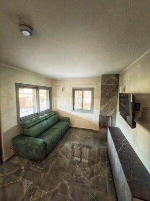 A room with a green sofa, two windows, a flat-screen TV, and a marble floor.