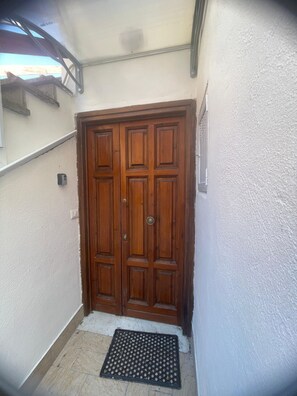A wooden double door with a metal handle, a small mat at the entrance, and a white textured wall.