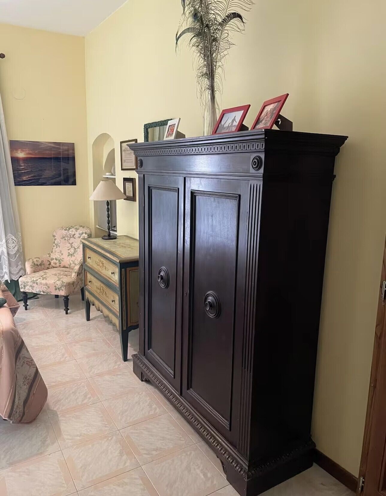 A room with a dark wooden cabinet, a chair, a lamp, and a painting on the wall.