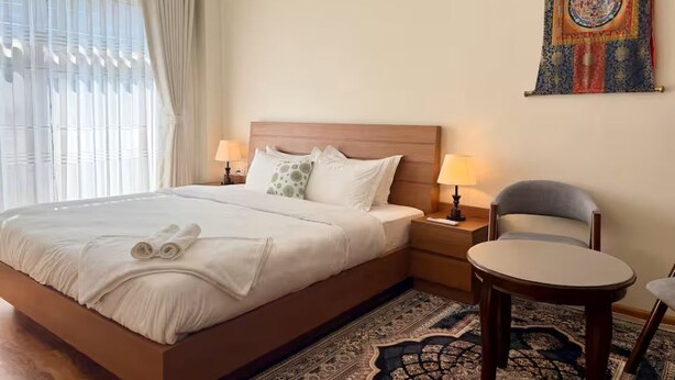Een hotelkamer met een bed, twee nachtkastjes met lampen, een stoel, een ronde tafel en een wandtapijt.