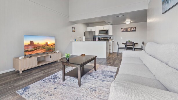 Ett modernt vardagsrum med en platt-TV, ett mörkt träbord, en soffa i ljusa färger och ett köksområde i bakgrunden.