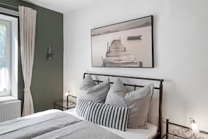 Un dormitorio con cama, almohadas, una mesita de noche, una lámpara de pared, una ventana con cortinas y una fotografía de un muelle.