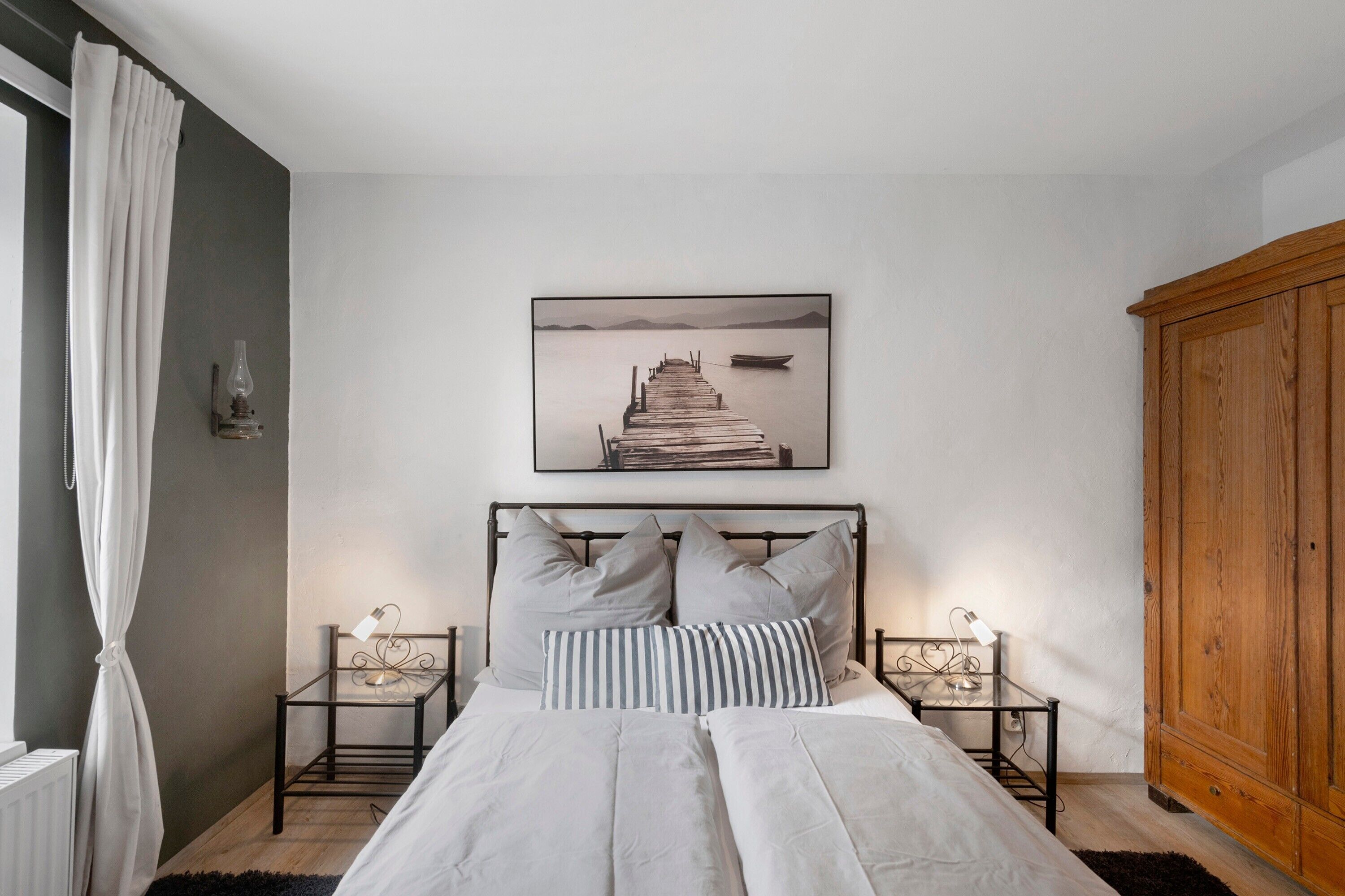 A bedroom with a bed, two bedside tables, a wooden wardrobe, a framed picture on the wall, and a window with curtains.