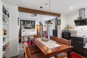Un comedor con mesa de madera, sillas y alfombra roja. Se observa una cocina con estufa, horno y alacenas. A la izquierda, se ve una estantería.