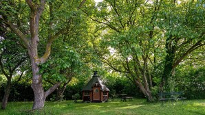 Une construction rustique en bois, dotée d’un toit de chaume, entourée d’une végétation luxuriante et d’arbres.